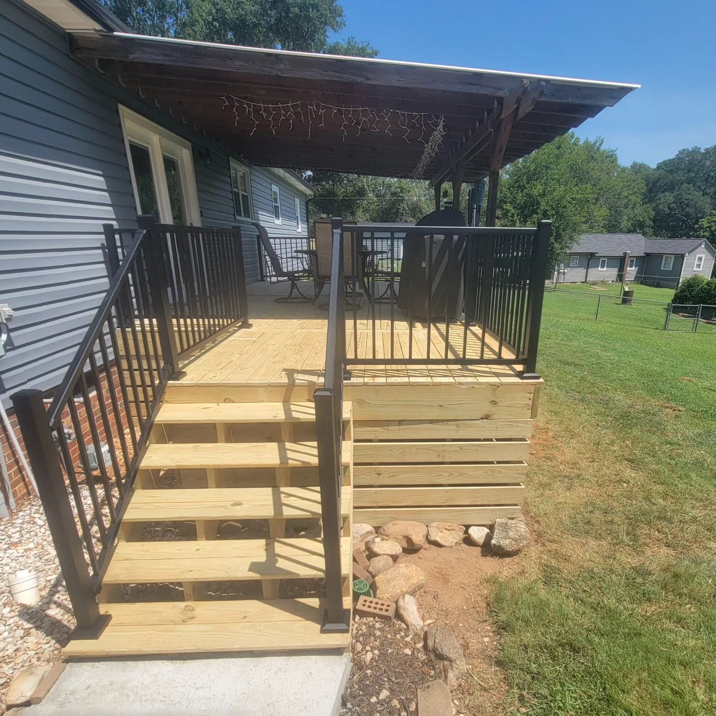 Wooden deck with black railing and pergola, steps lead to the deck, next to a house with blue siding.
