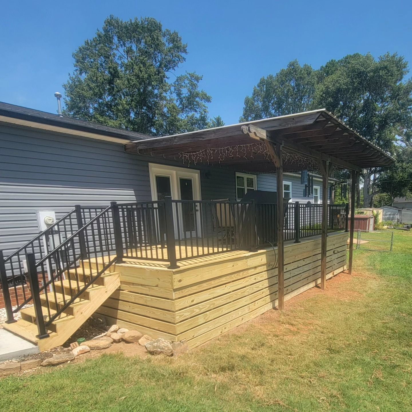 Wooden deck with black railings and a metal awning attached to a gray house, set on green grass.