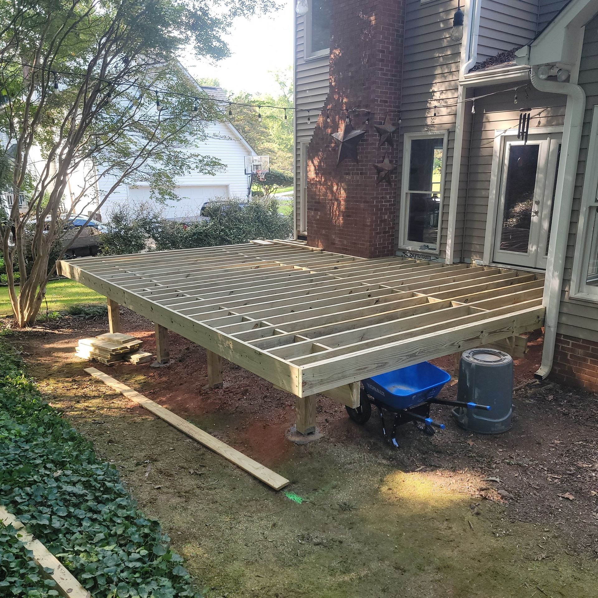 Newly constructed wooden deck attached to a house with a brick chimney. A blue wheelbarrow and barrel are under the deck.
