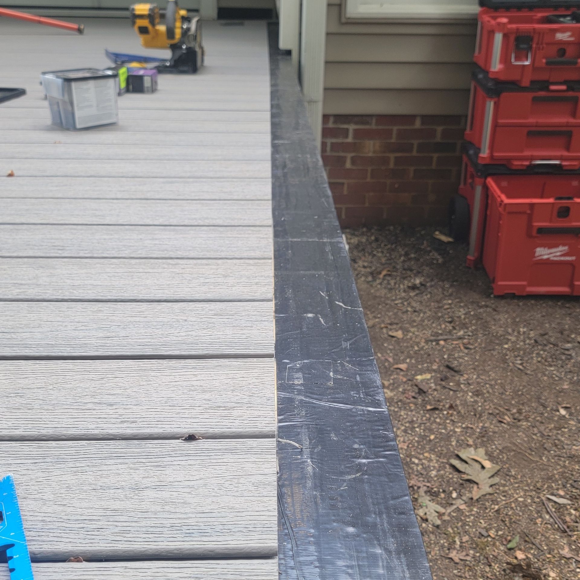 A deck with gray composite boards next to a black timber border, beside a brick wall and dirt.
