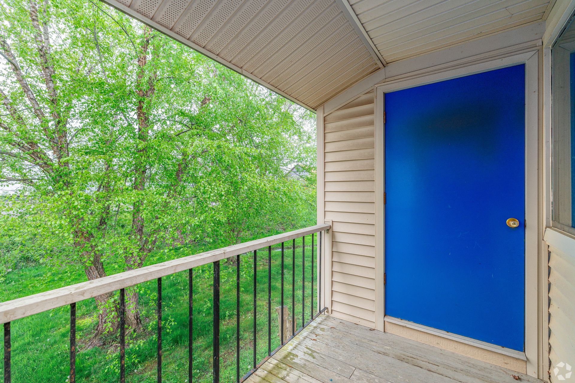 Apartment balcony with blue door, beige siding, and green trees beyond the railing
