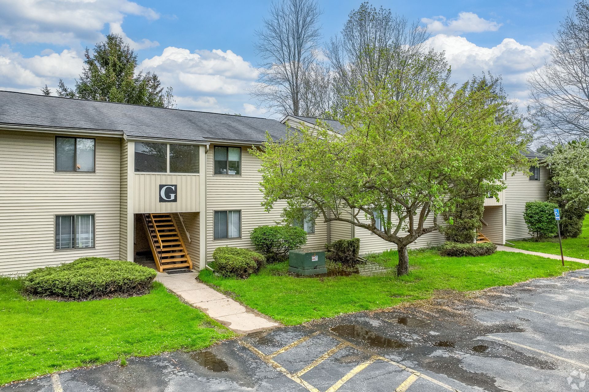 Apartment building exterior with green lawn, trees, and a parking lot under a cloudy sky