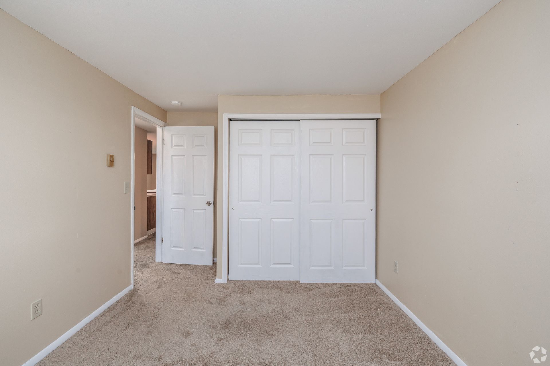 Empty beige bedroom with carpet, white closet doors, and an open doorway to a hall
