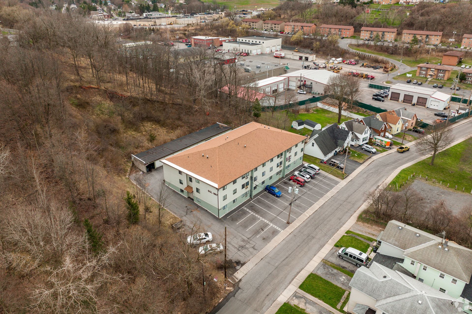 Aerial view of an apartment building beside a road, with trees and homes in a suburban neighborhood.