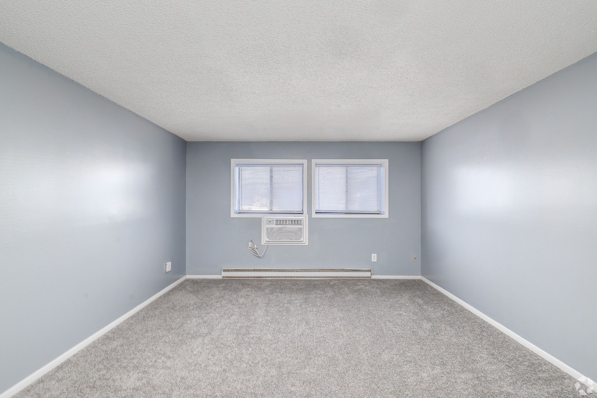 Empty light-blue room with gray carpet, two windows, and a wall-mounted AC unit