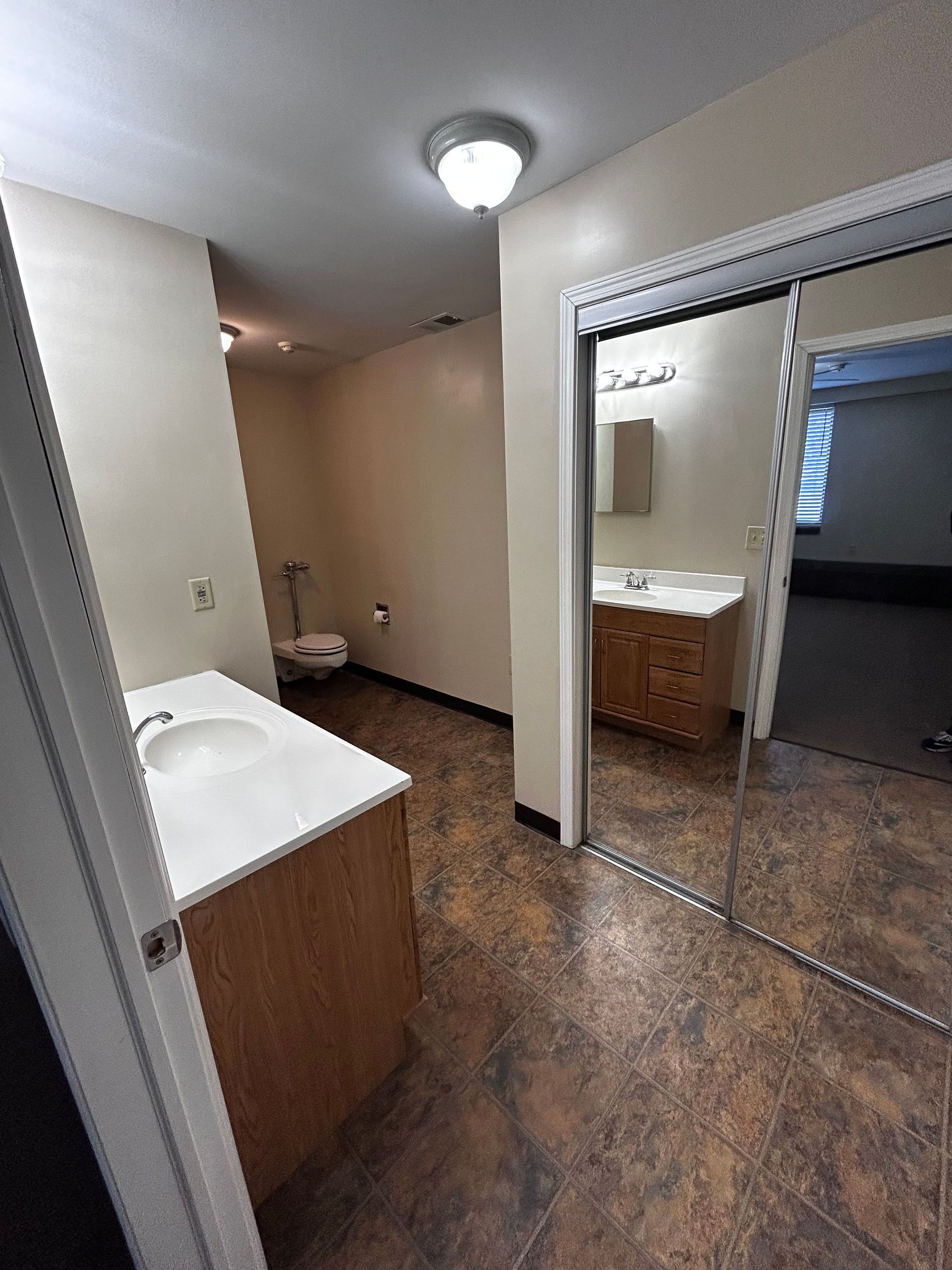 Small bathroom with sink, mirrored closet doors, and brown tile flooring; hallway visible beyond.