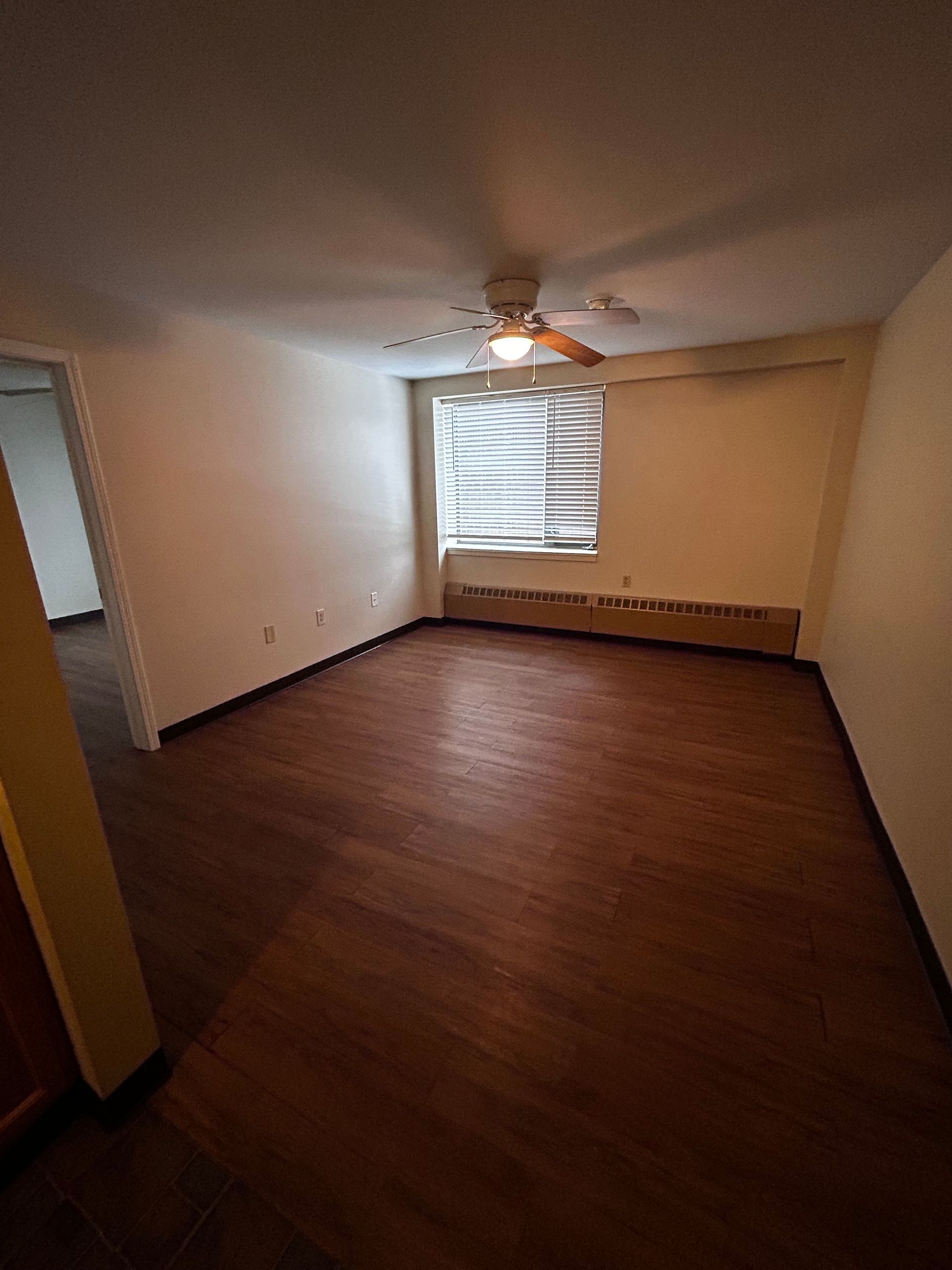 Empty room with brown wood floor, beige walls, ceiling fan, and a window with blinds.