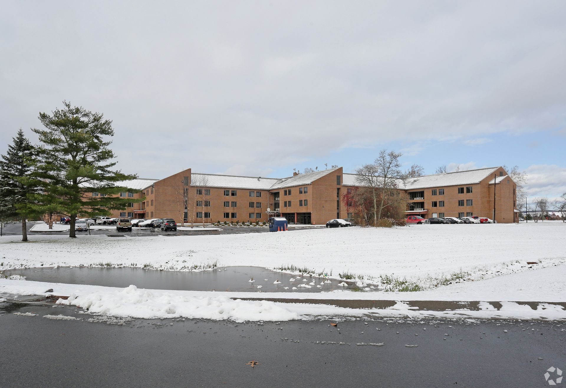 Snow-covered brick apartment building with parked cars under a cloudy winter sky