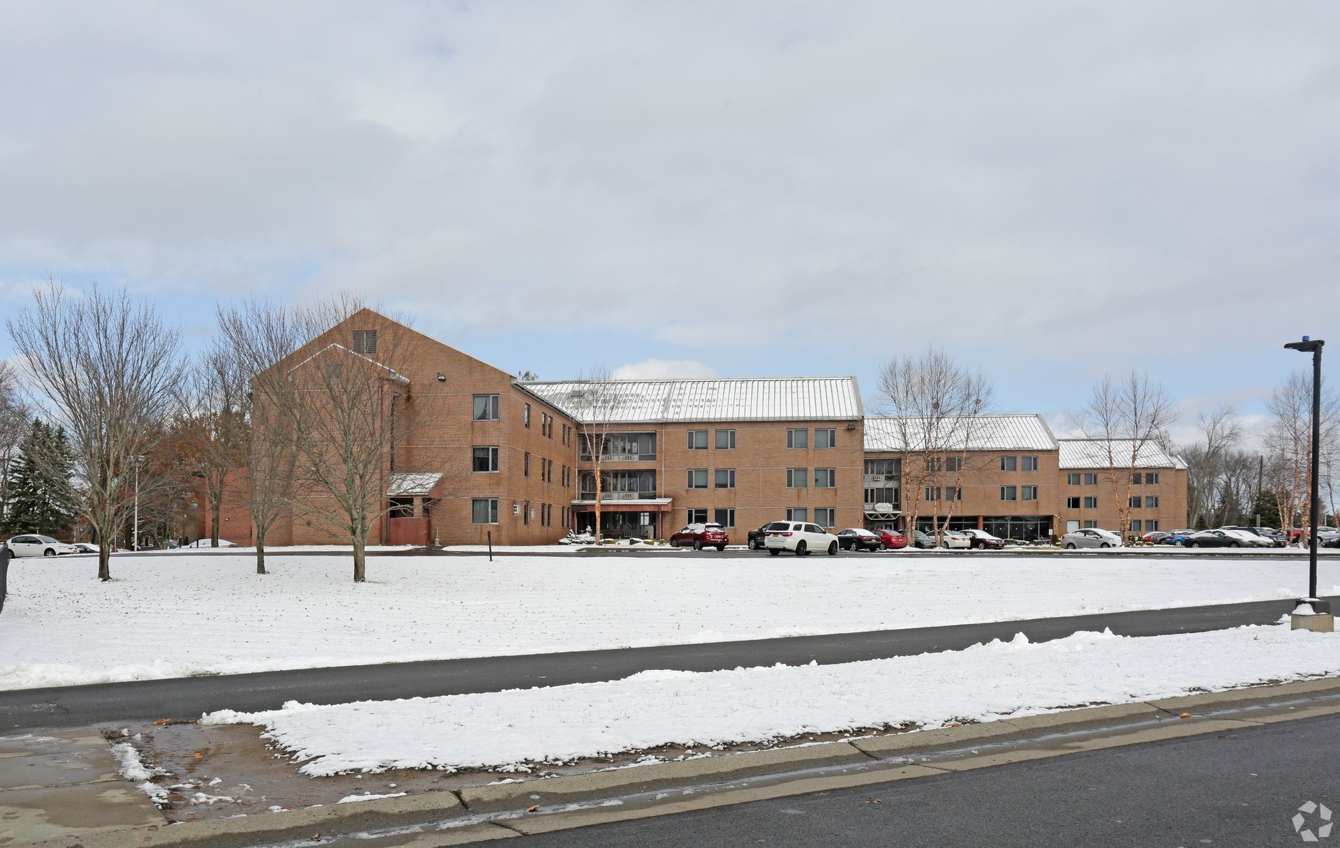 Snowy brick apartment building with parked cars along a road under an overcast sky