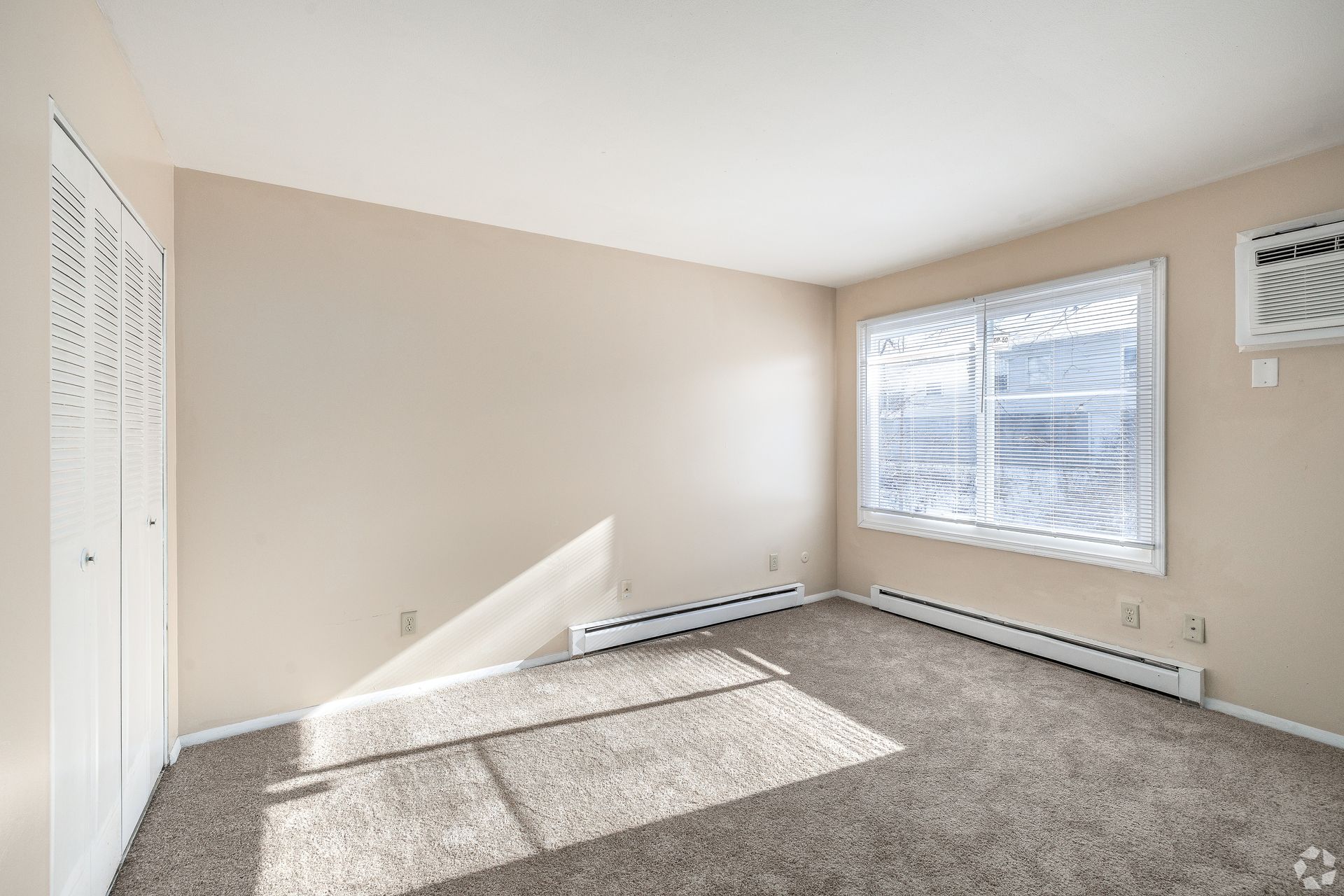 Empty beige bedroom with carpet, white window blinds, and sunlight on the floor