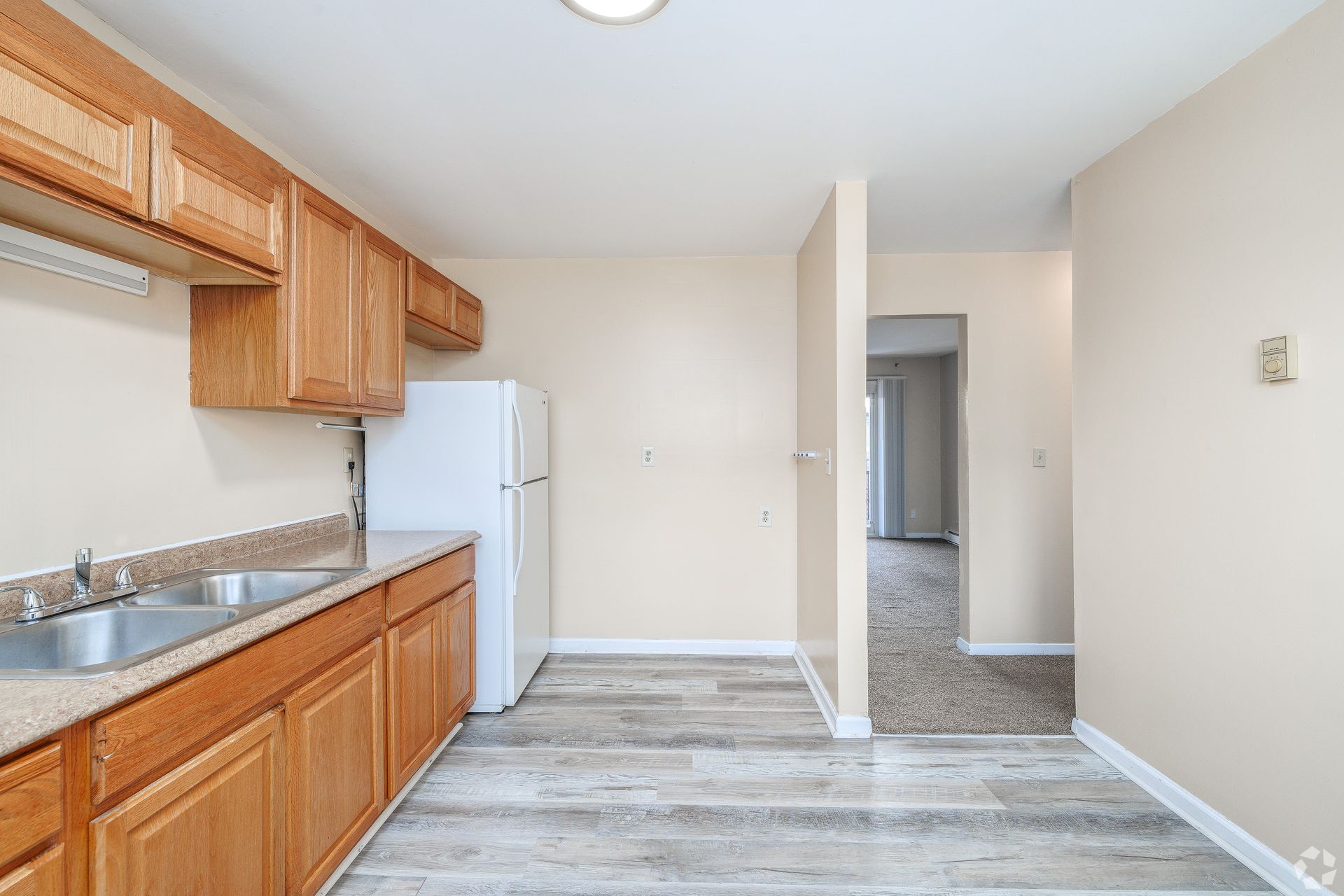 Empty kitchen with wood cabinets, white refrigerator, and light gray flooring leading to a hallway.