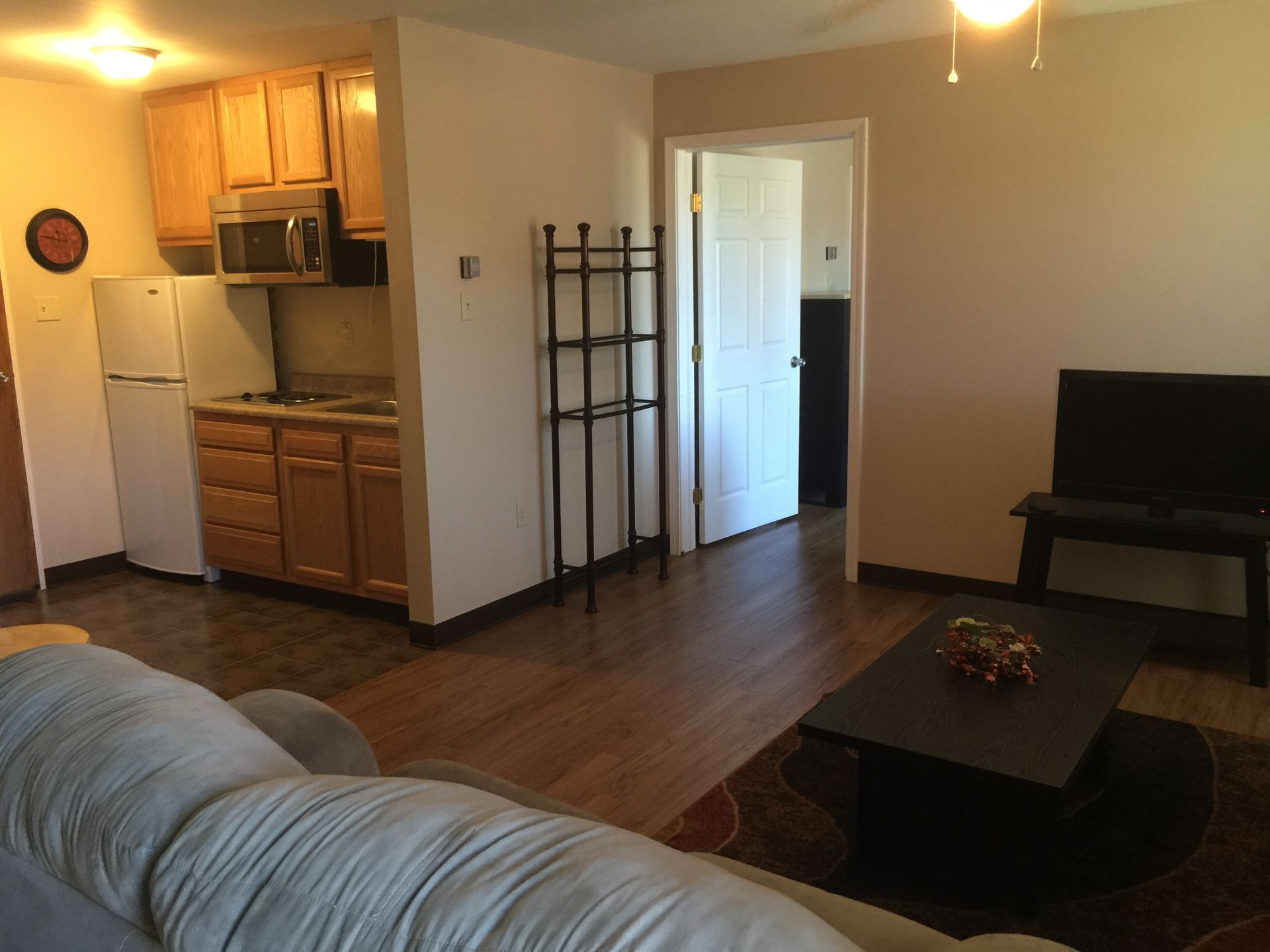 Open-plan living room with kitchen, hardwood floors, gray sofa, coffee table, and a piano by the wall.