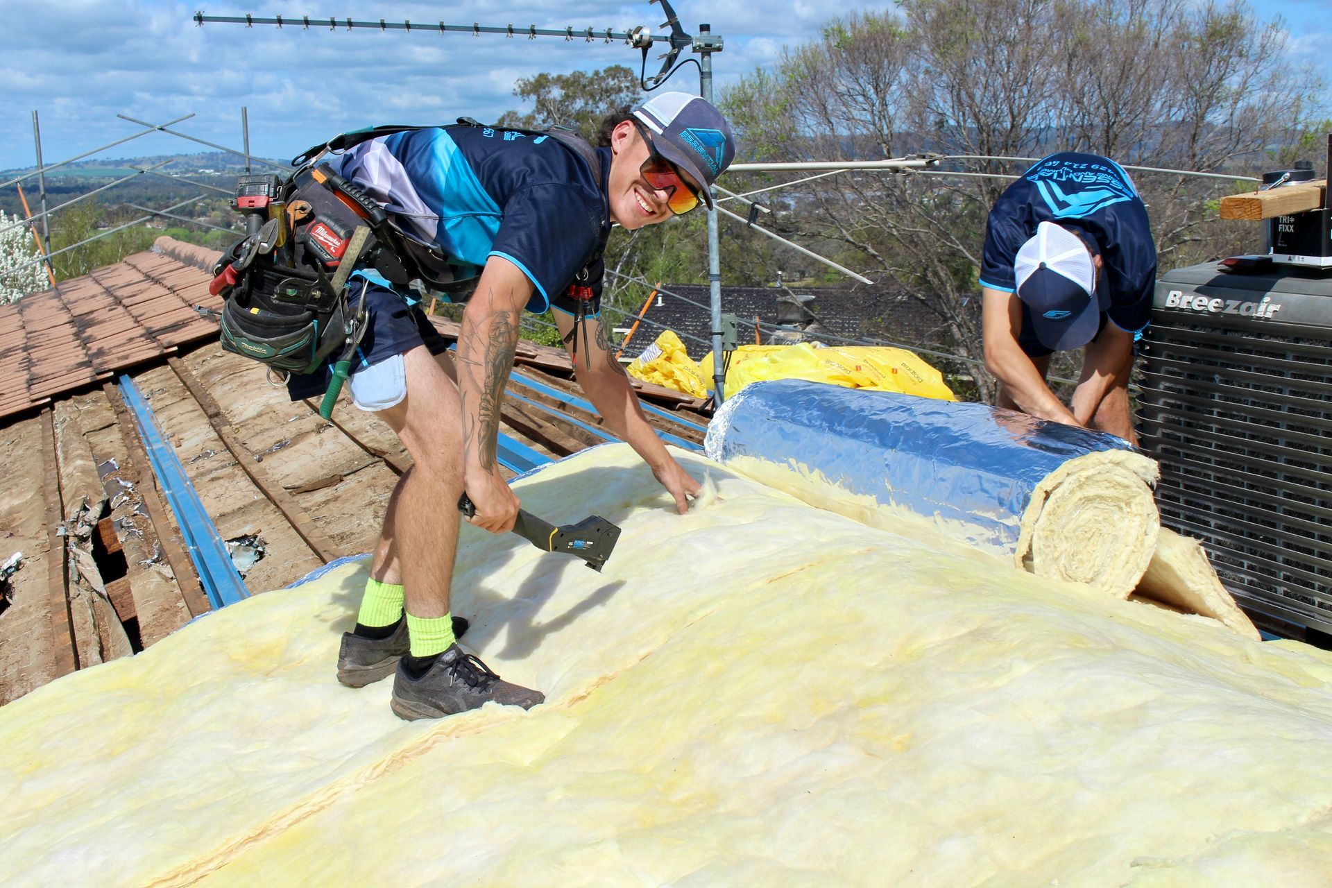 Two men working on roof installation with one smiling at the camera — Essential Roofing and Plumbing in Wagga Wagga, NSW