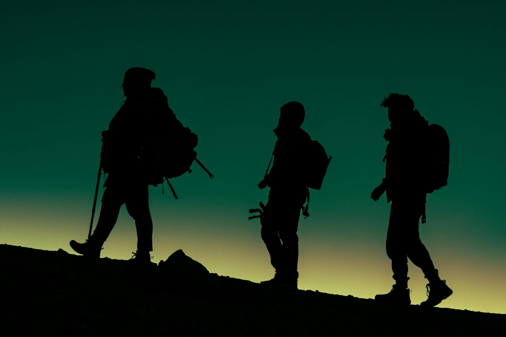 Three hikers in silhouette on a ridge at dusk under a green-blue sky