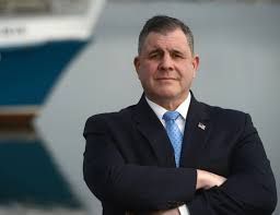 Man in suit, arms crossed, stands in front of a cruise ship.