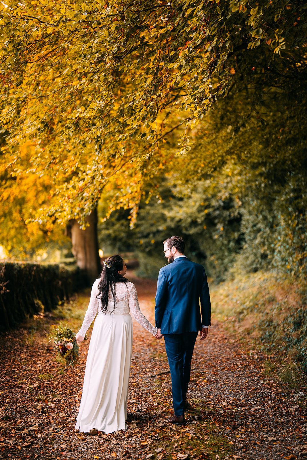 A bride and groom are walking down a path holding hands.
