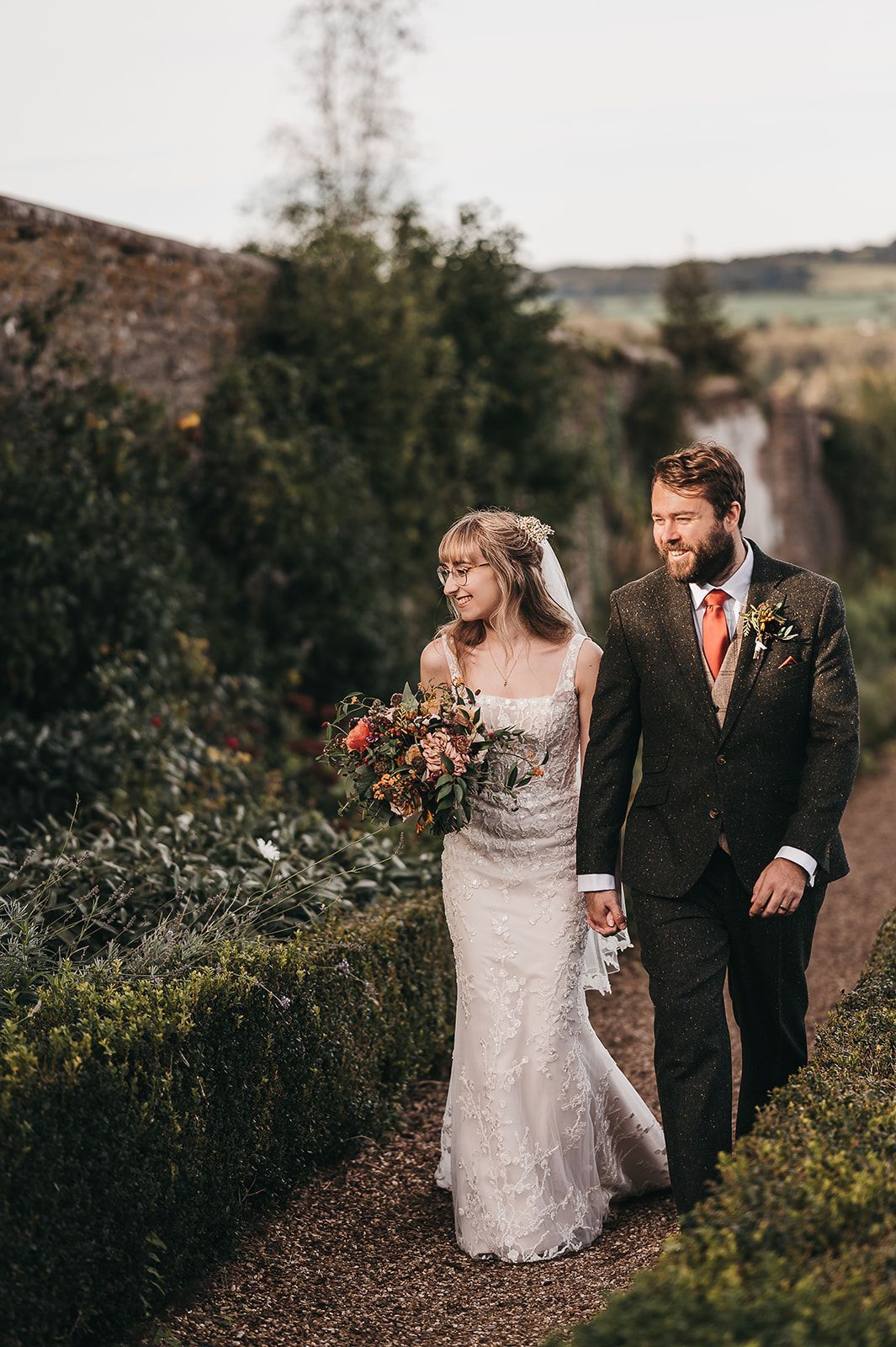 A bride and groom are walking down a path holding hands.