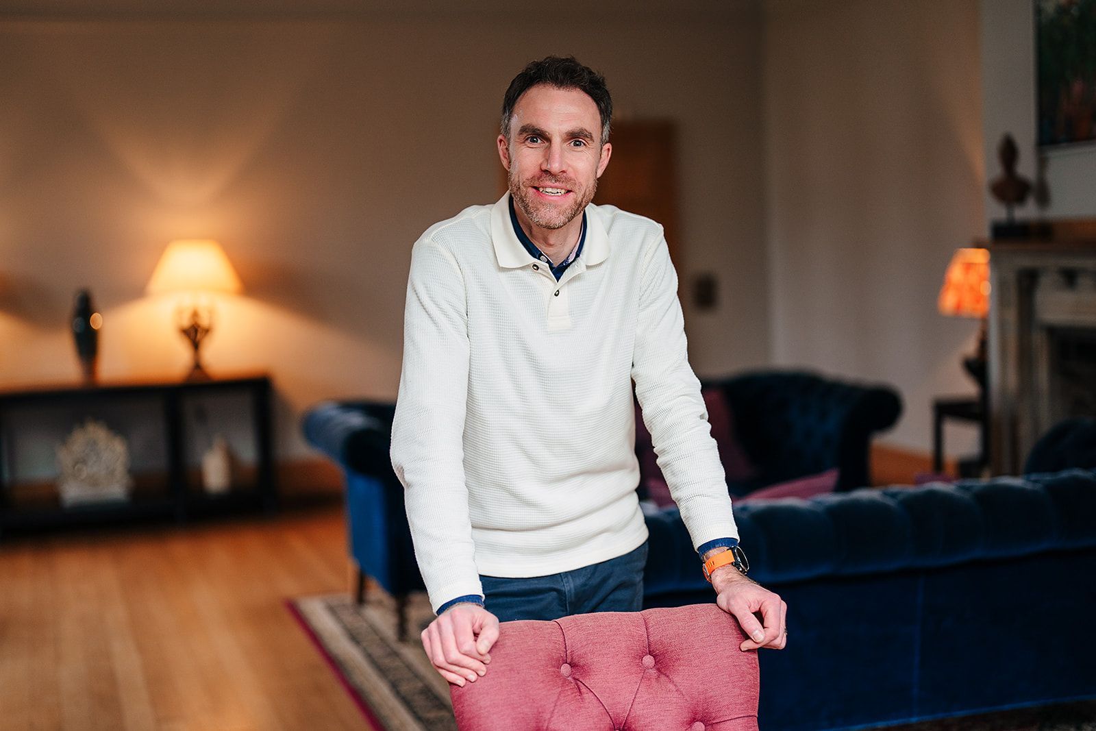 A man in a white sweater is standing next to a pink chair in a living room.