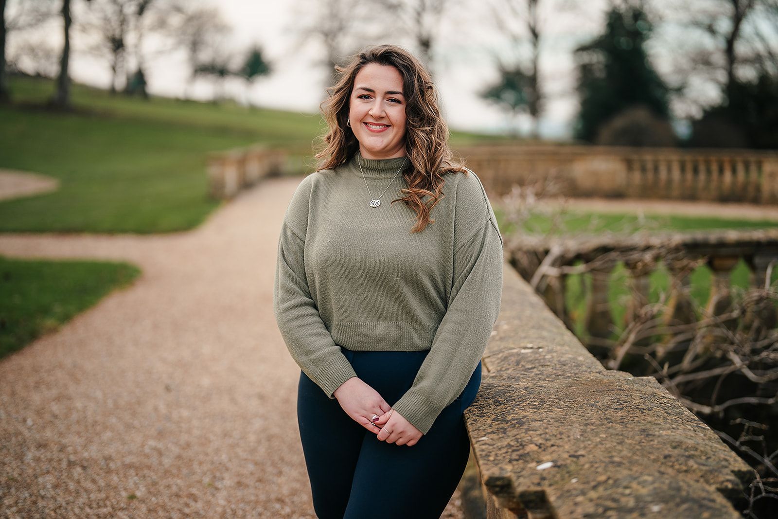 A woman in a green sweater is standing next to a stone wall in a park.