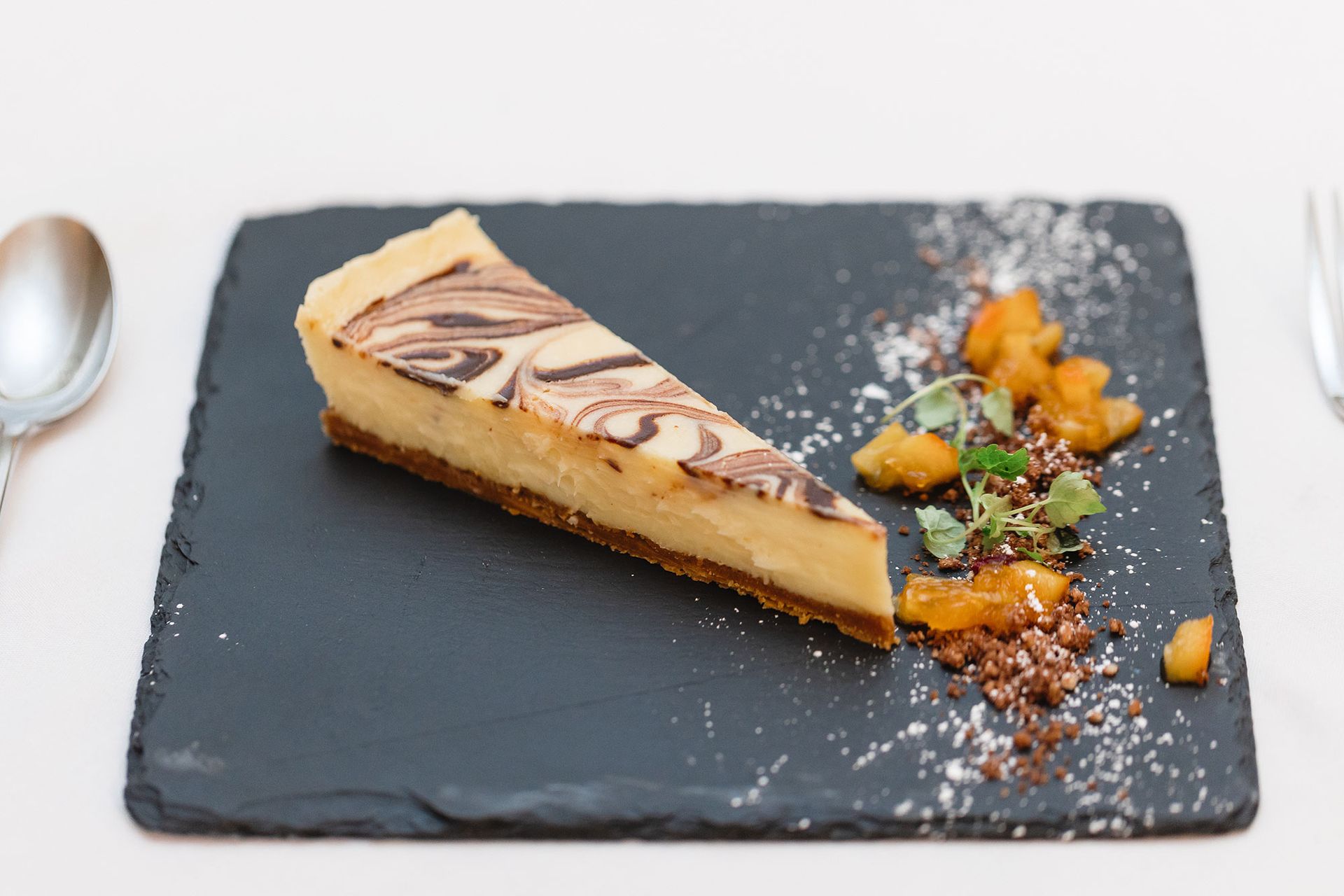 A slice of cheesecake on a black plate with a fork and spoon