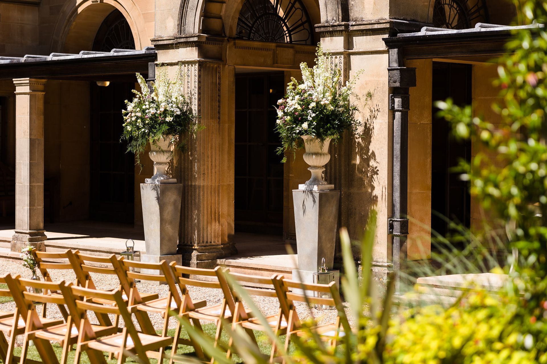 A row of wooden chairs are lined up in front of a building with flowers in vases.