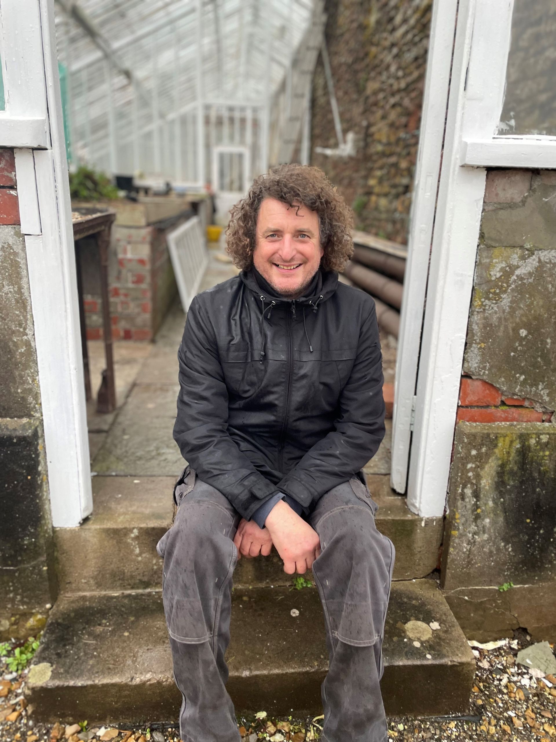 A man with curly hair is sitting on the steps of a greenhouse.