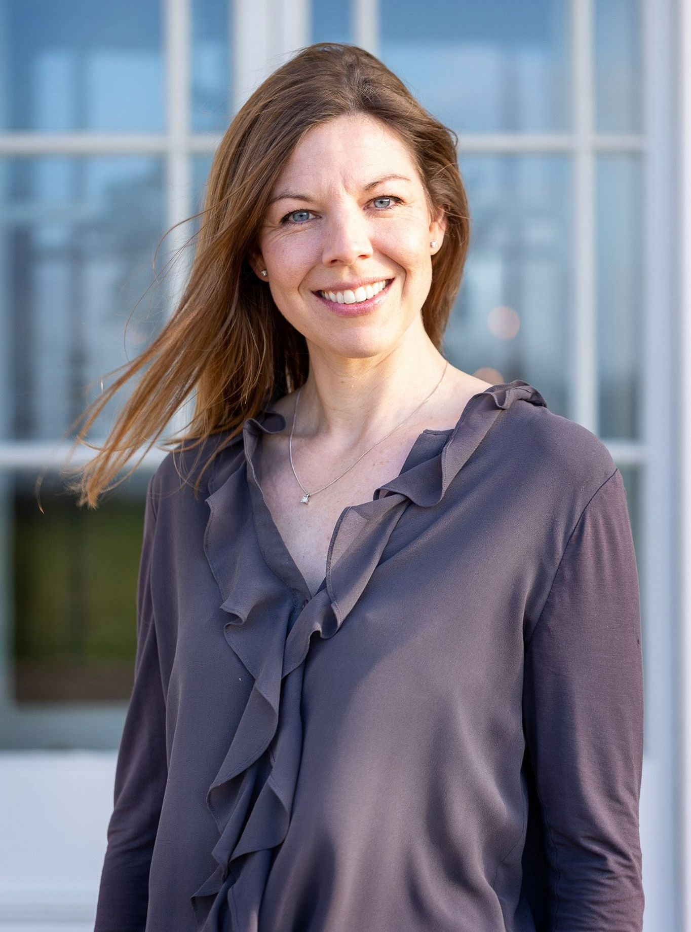 A woman in a ruffled shirt is smiling in front of a window.