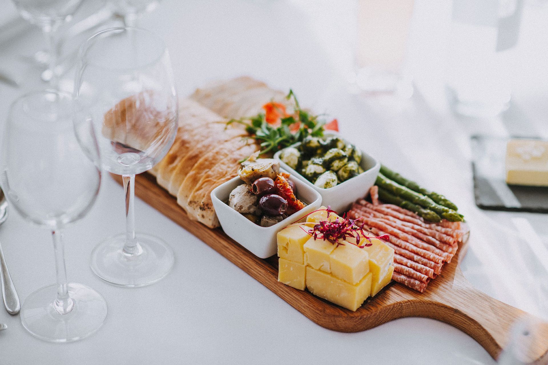A wooden cutting board filled with food and wine glasses on a table.