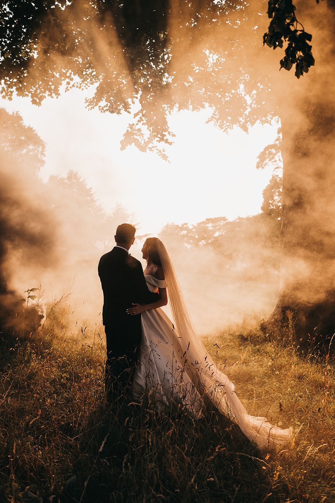 A bride and groom are standing in a field with smoke coming out of the trees.