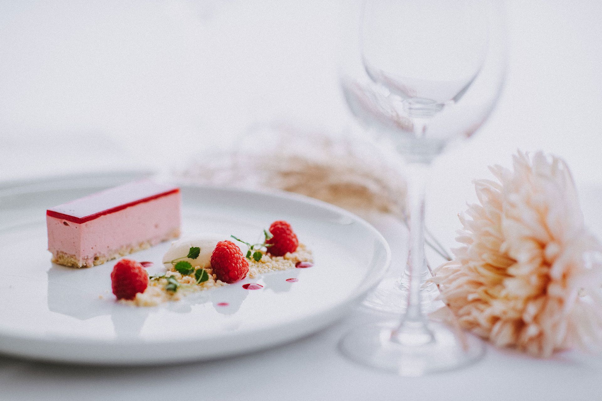 A white plate topped with a piece of cake and raspberries next to a wine glass.