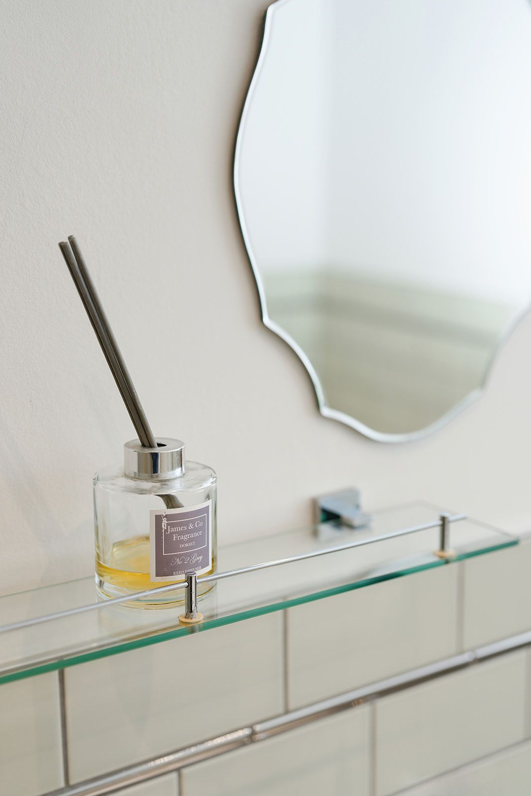 A bottle of reed diffuser is sitting on a glass shelf in a bathroom next to a mirror.