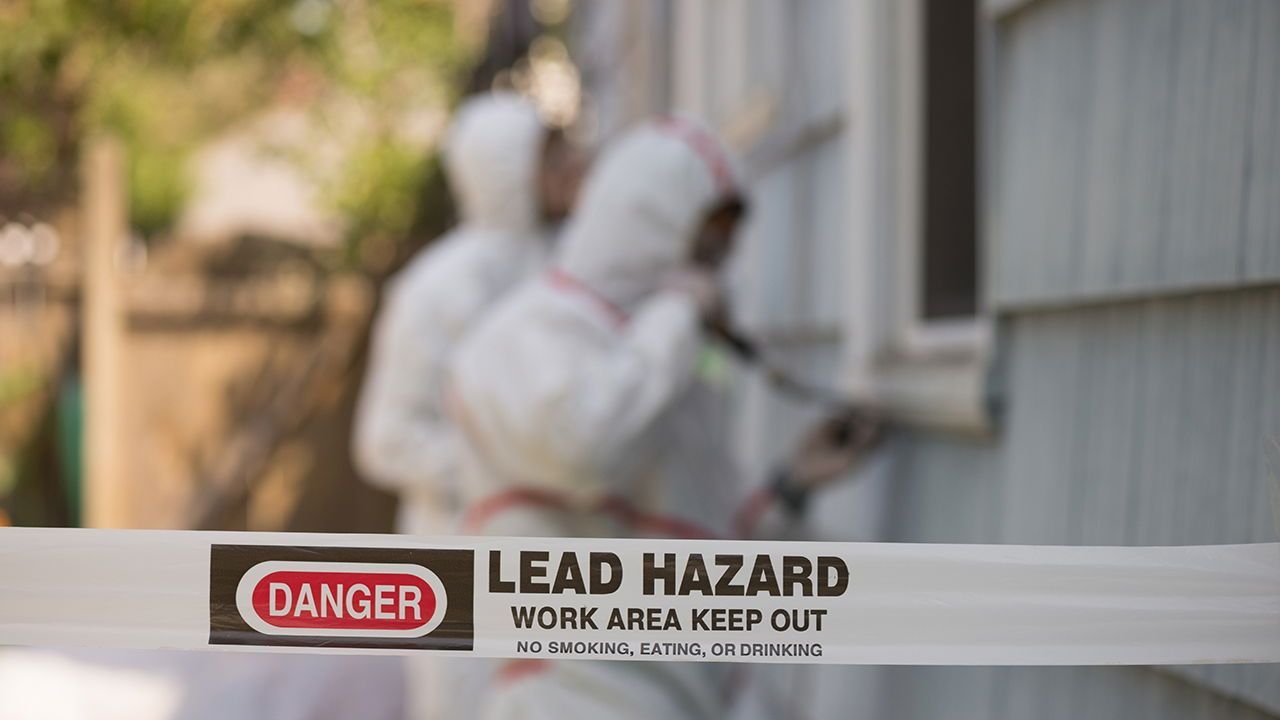 Two people in protective suits work on a house exterior behind a 