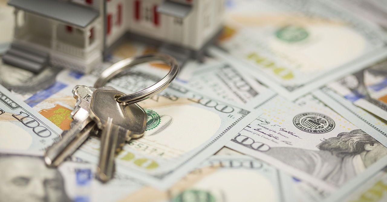Keys and a miniature house sit on a pile of $100 bills, suggesting a real estate purchase.