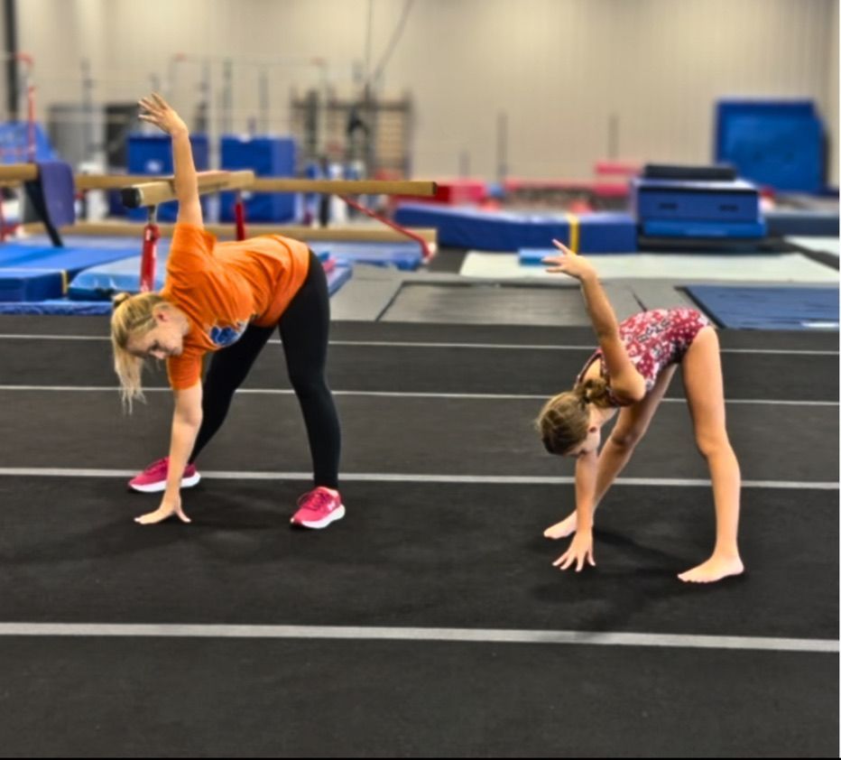 Woman and girl doing a yoga pose, arm raised, hand touching toes, in a gym.