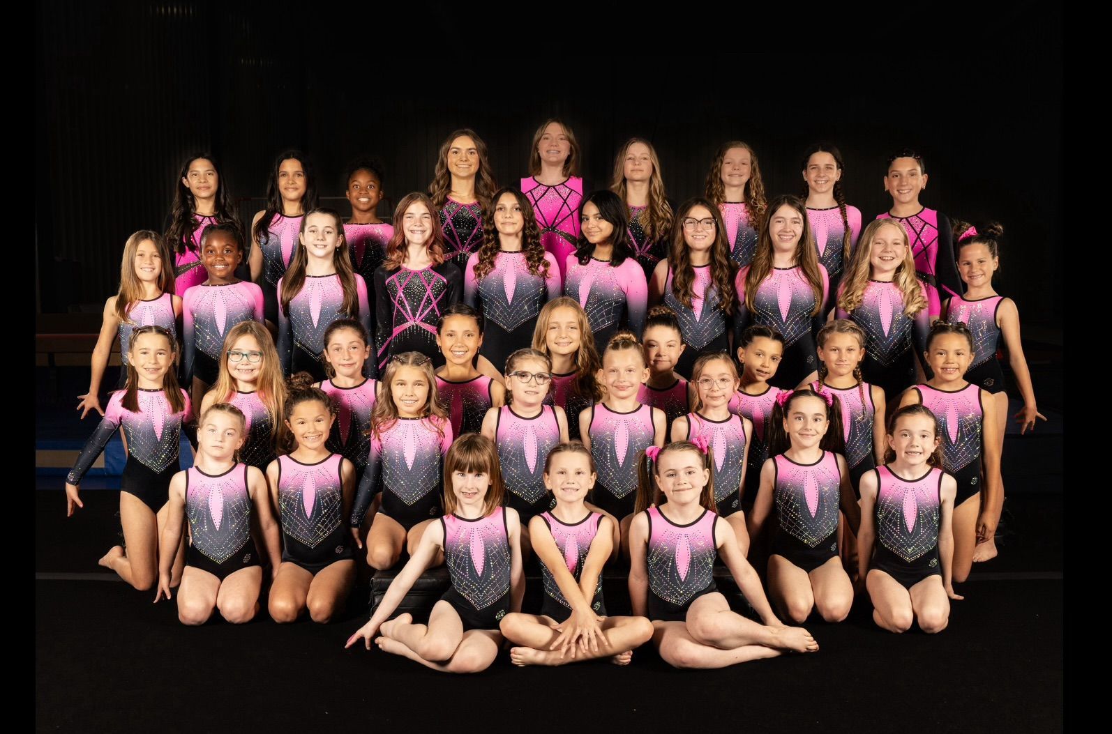 Group photo of young gymnasts in pink and black leotards, posing against a black backdrop.