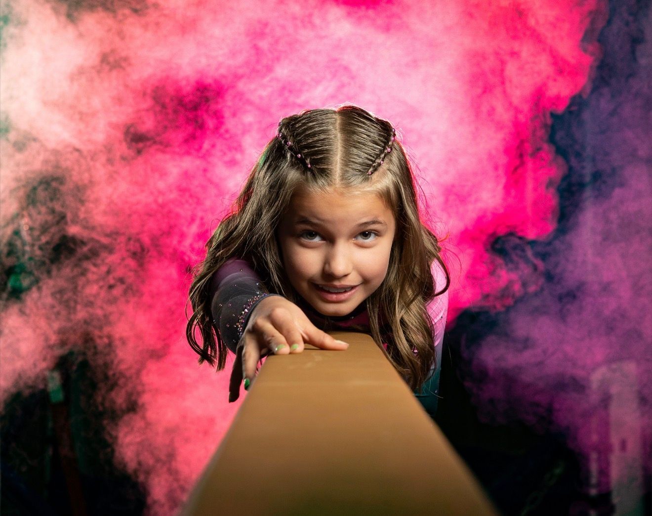 Girl on balance beam, reaching forward with pink and purple smoke background.