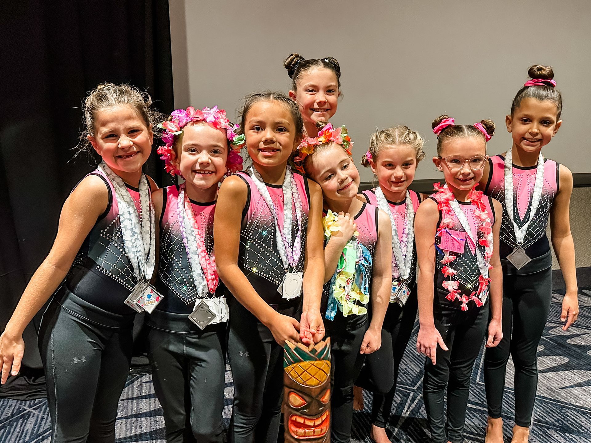 Group of young dancers in matching outfits pose with medals and a tiki.