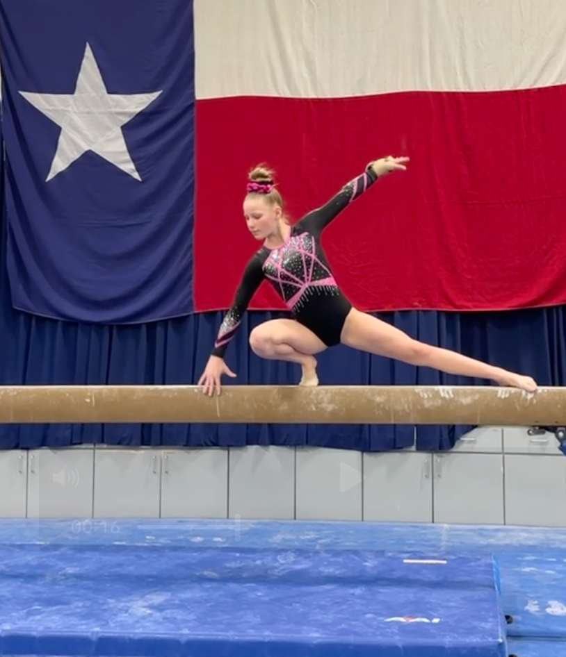 Gymnast in a pink and black leotard balances on a beam, arms raised, dark background.