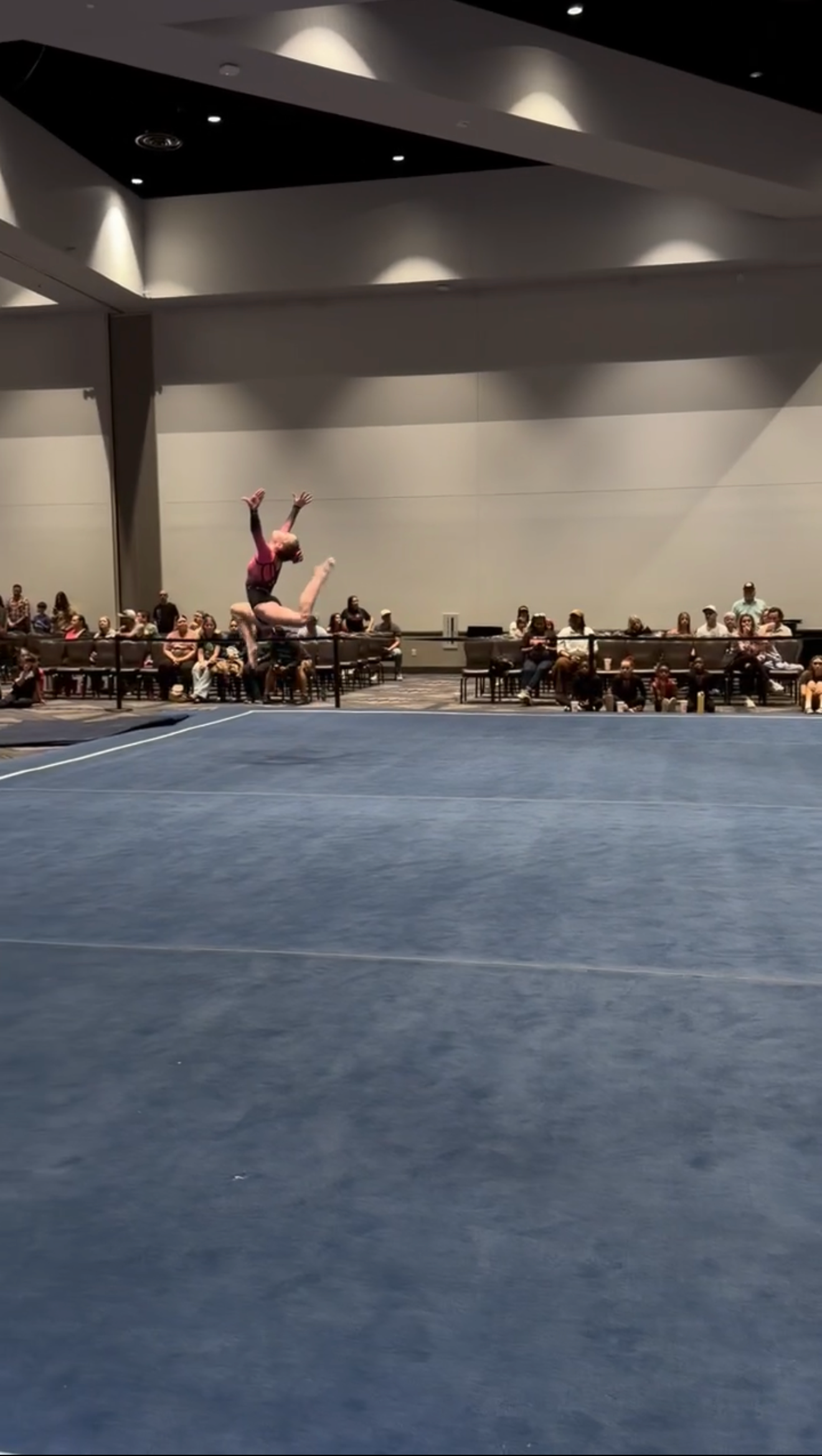 Gymnast in mid-air performing a flip on a blue mat, audience in the background.