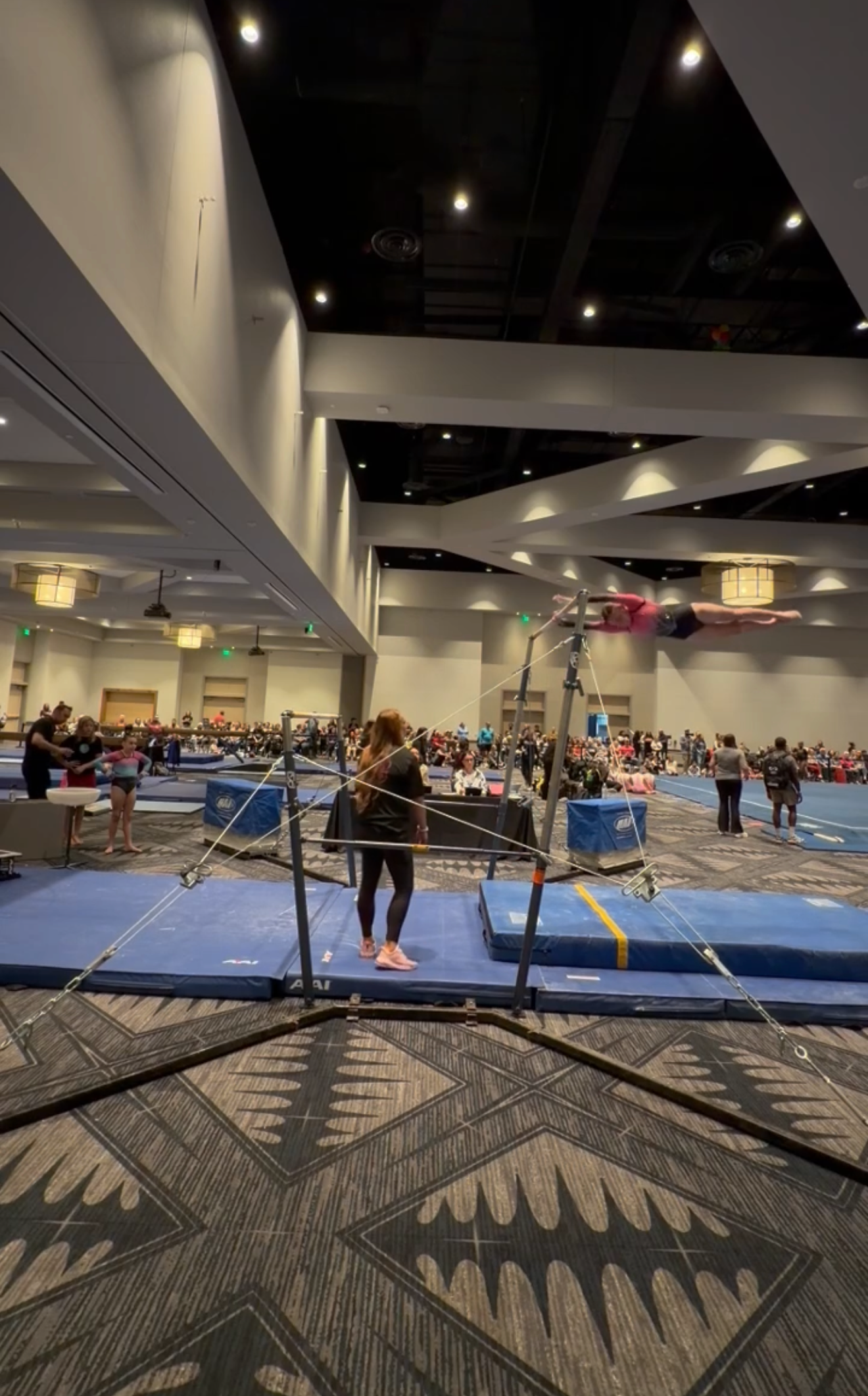 Gymnast standing near uneven bars in a large indoor competition setting with blue mats and spectators.
