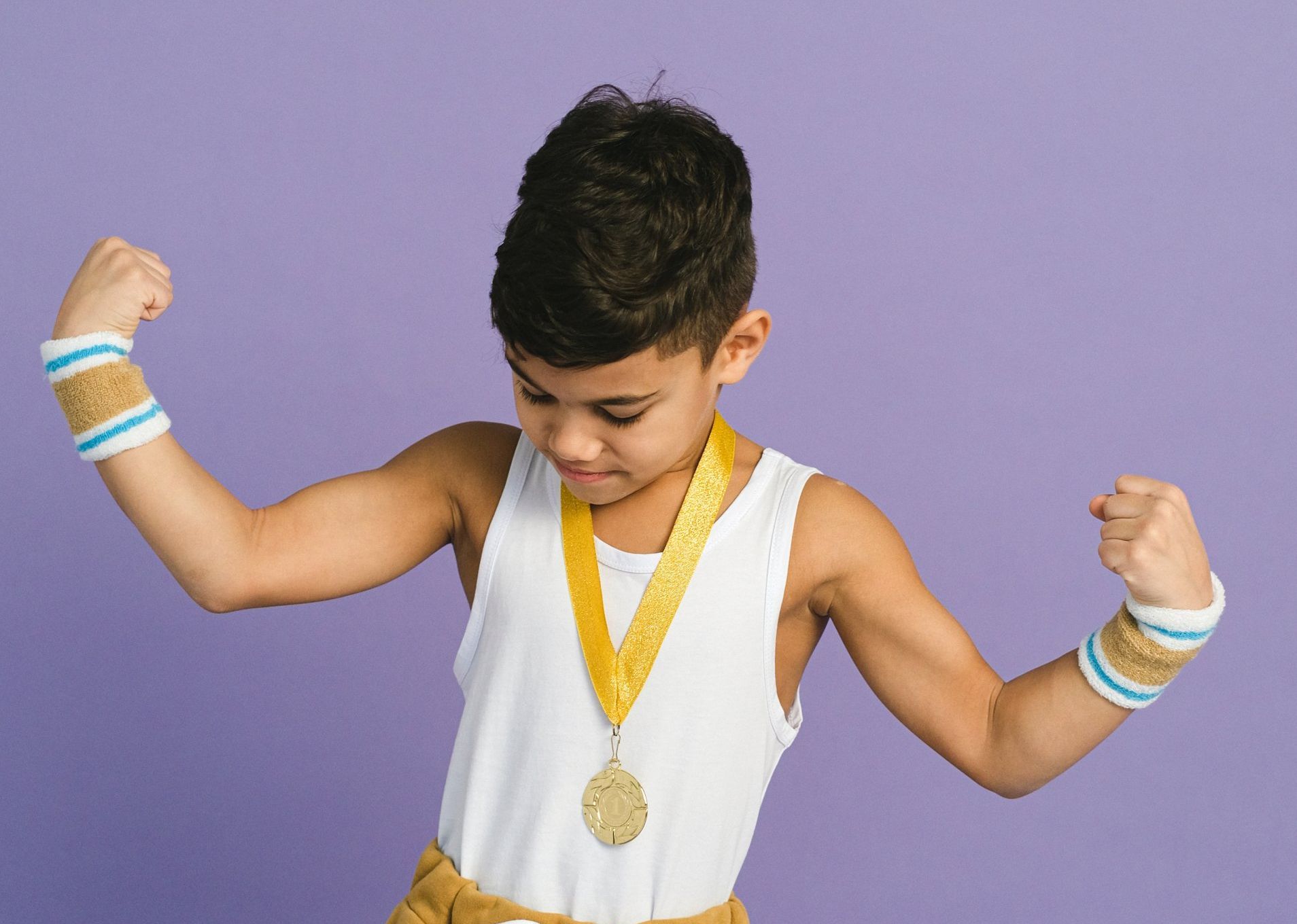 Boy flexing arms, wearing a medal, white tank top, and wristbands against a purple background.
