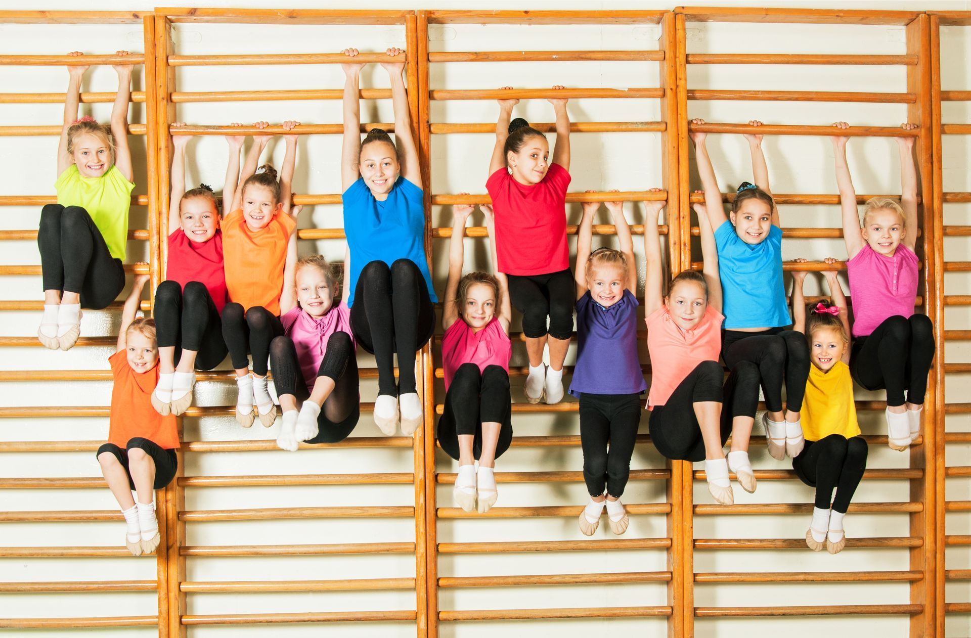 Girls in colorful shirts hanging from wooden wall bars.