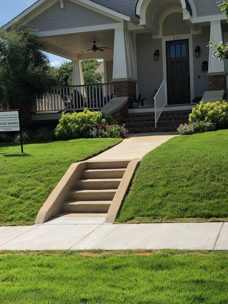 A concrete pathway and steps lead to a light gray house with a porch and dark brown door.