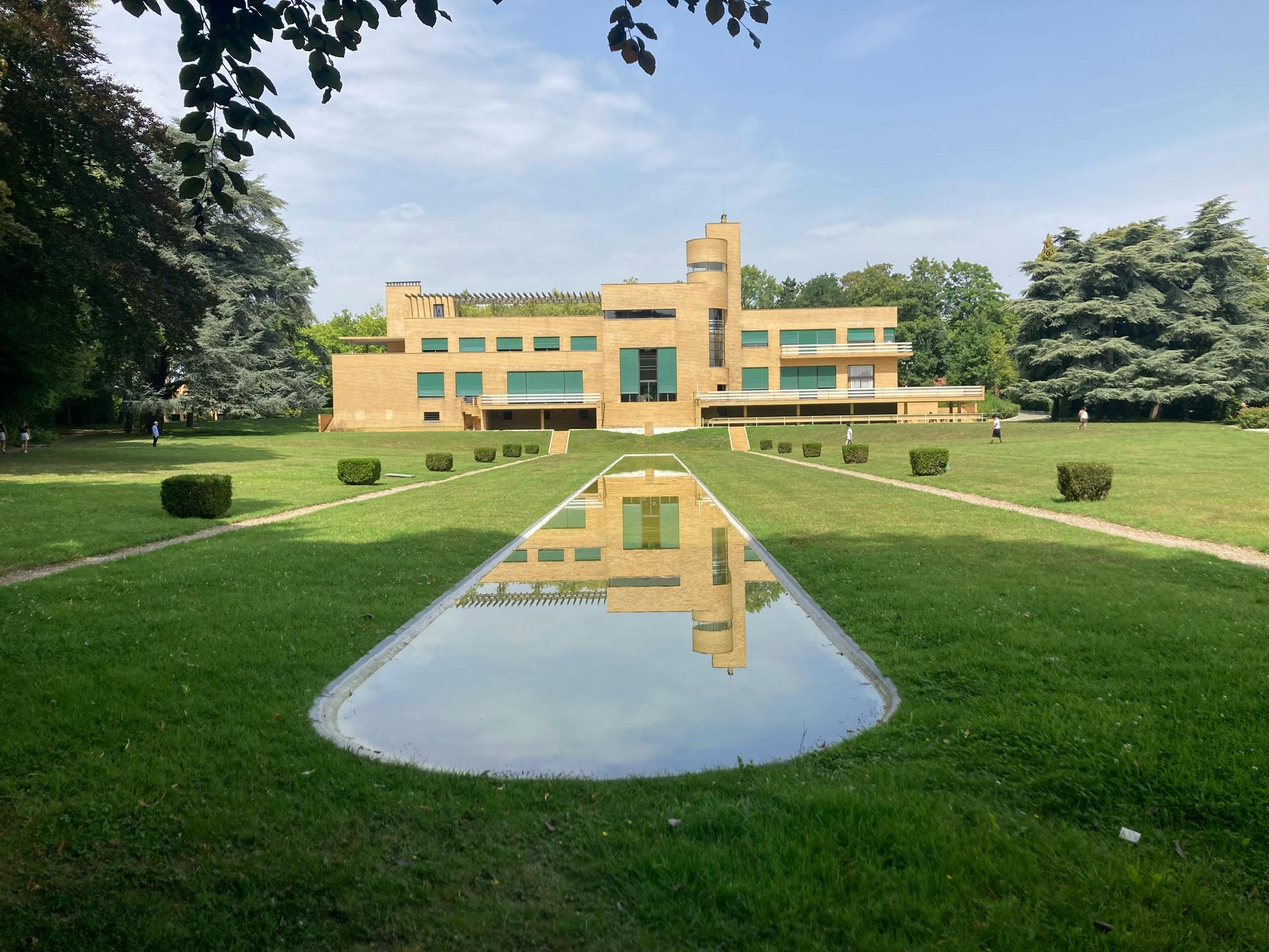 Yellow modernist building with a long reflecting pool on a green lawn, trees in background.