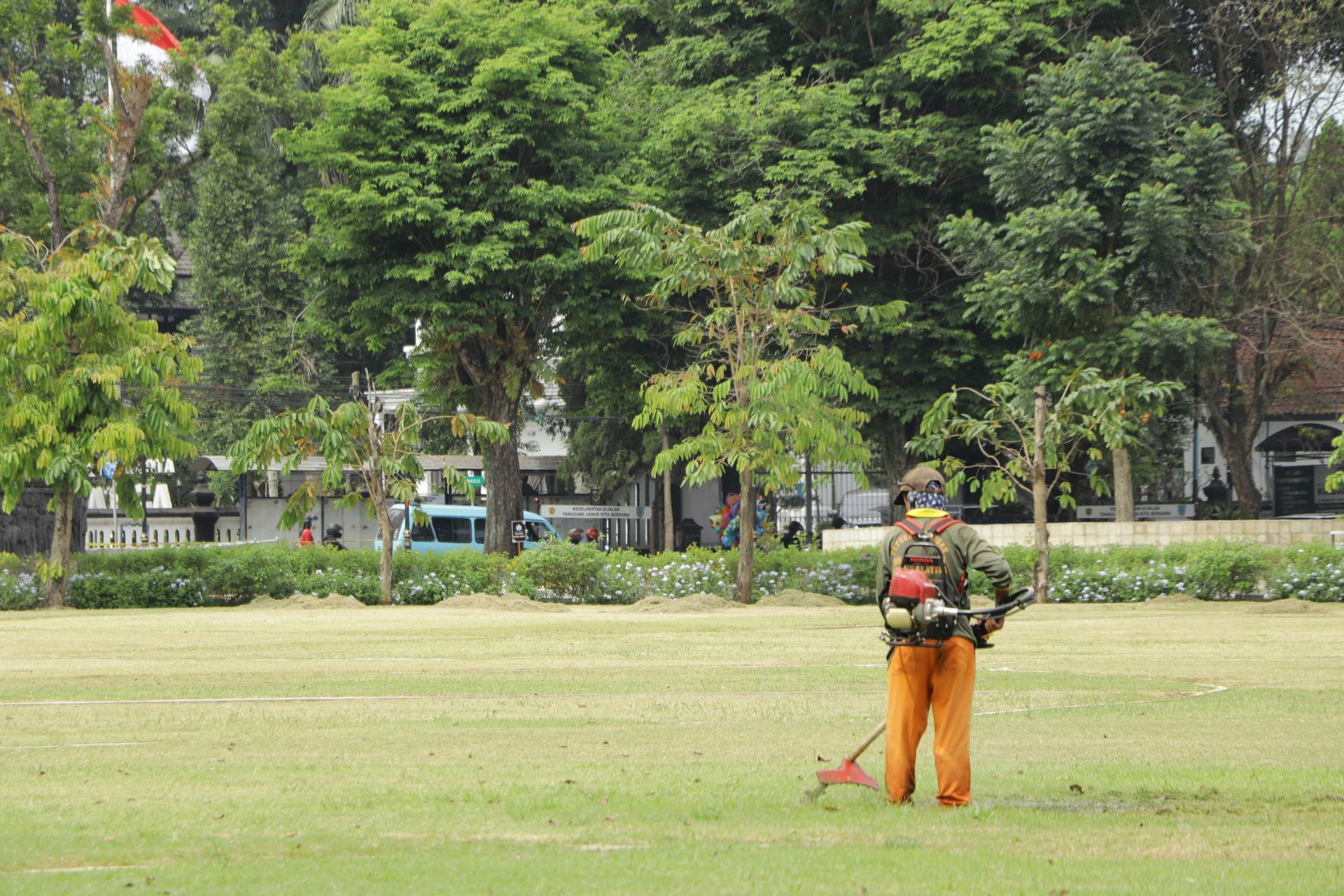 Man in orange pants and backpack weed-whacking a grassy field with trees and buildings in the background.