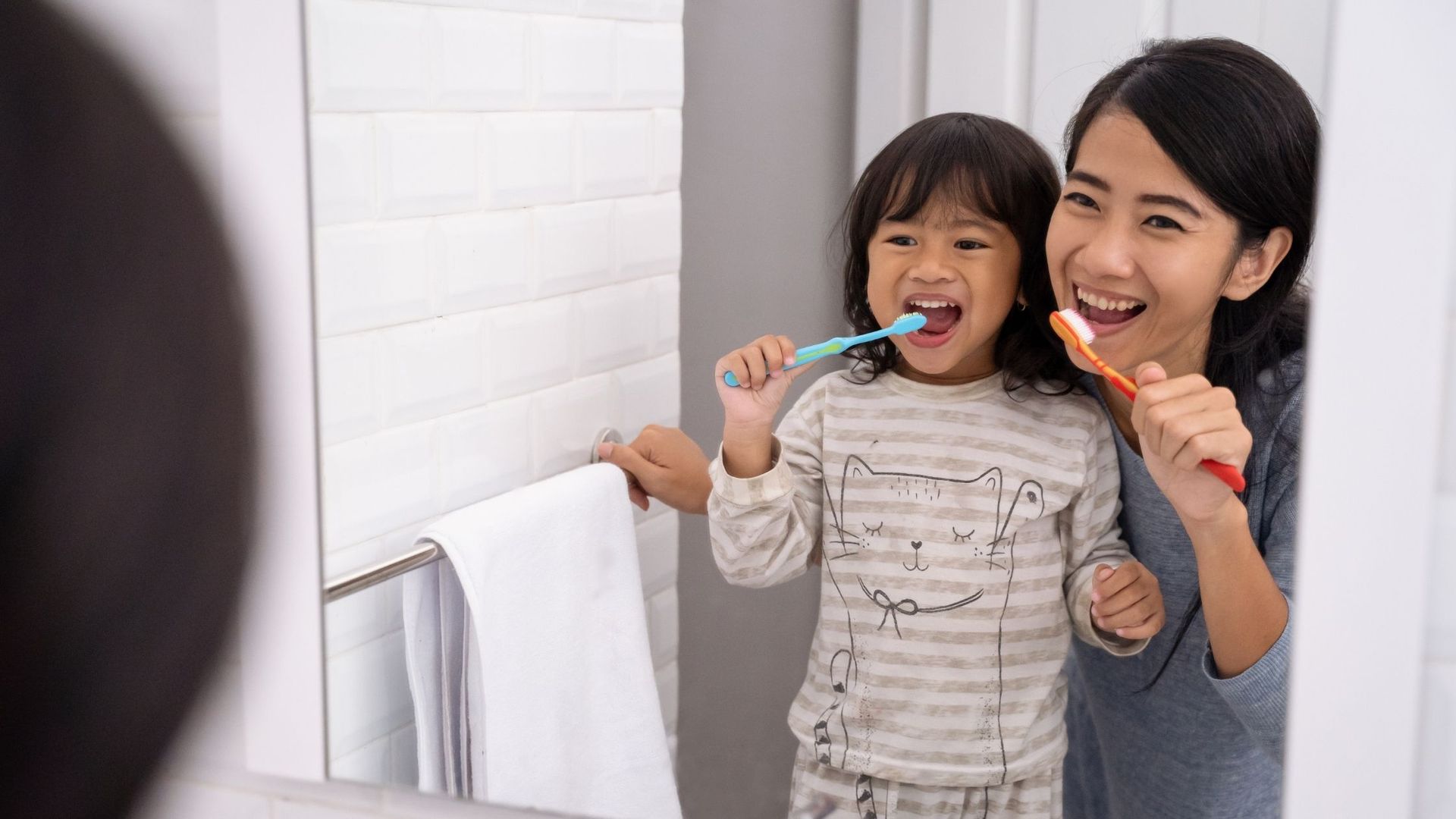 Woman and child brushing teeth together in a bathroom, smiling with toothbrushes.
