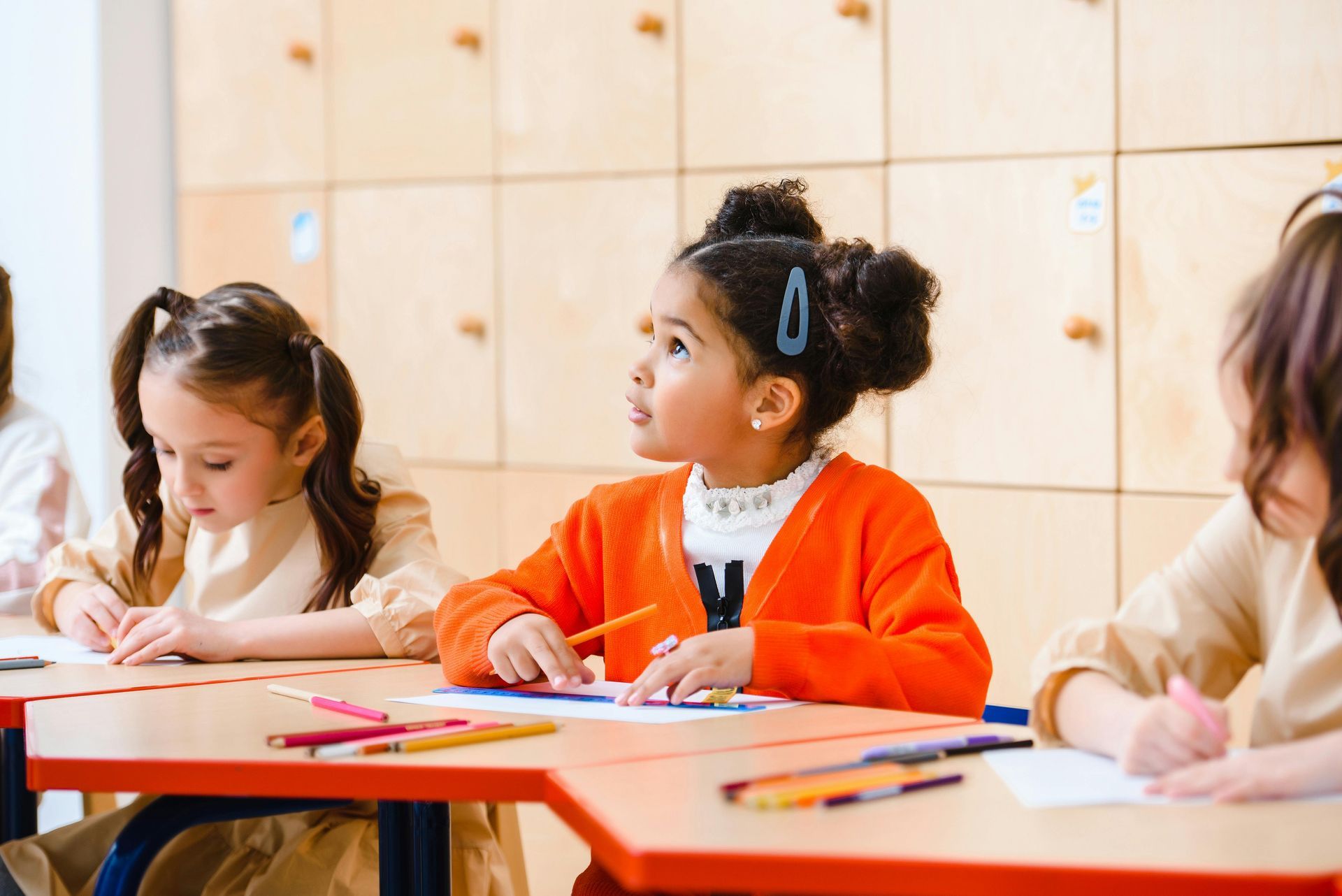 Children at desks in a classroom, drawing with crayons. One girl in orange cardigan looks up.