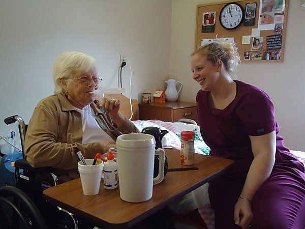 An elderly woman in a wheelchair is sitting at a table with a nurse.