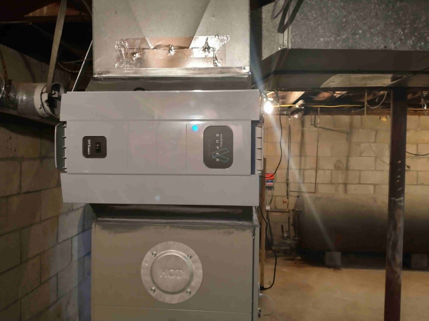 A light gray electronic air cleaner mounted on a furnace duct system in a basement with a concrete block wall background.