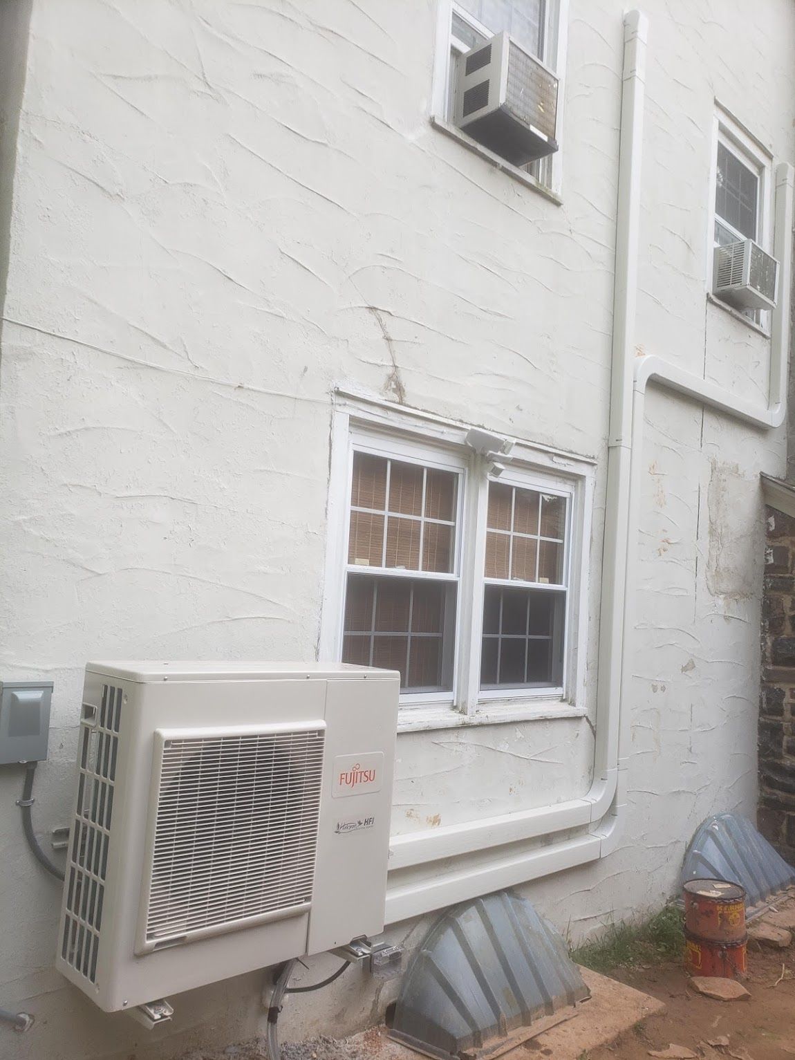 Exterior of a white stucco building with a mini-split AC unit, window air conditioners, and a ground-level window well.